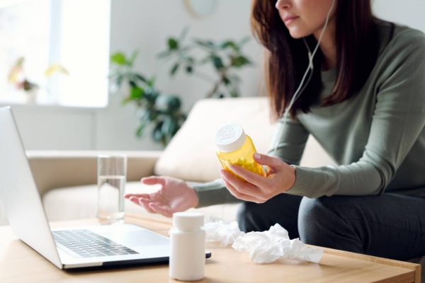 Sick female in homewear holding bottle of pills while sitting on couch by table in front of laptop at home and consulting with online doctor